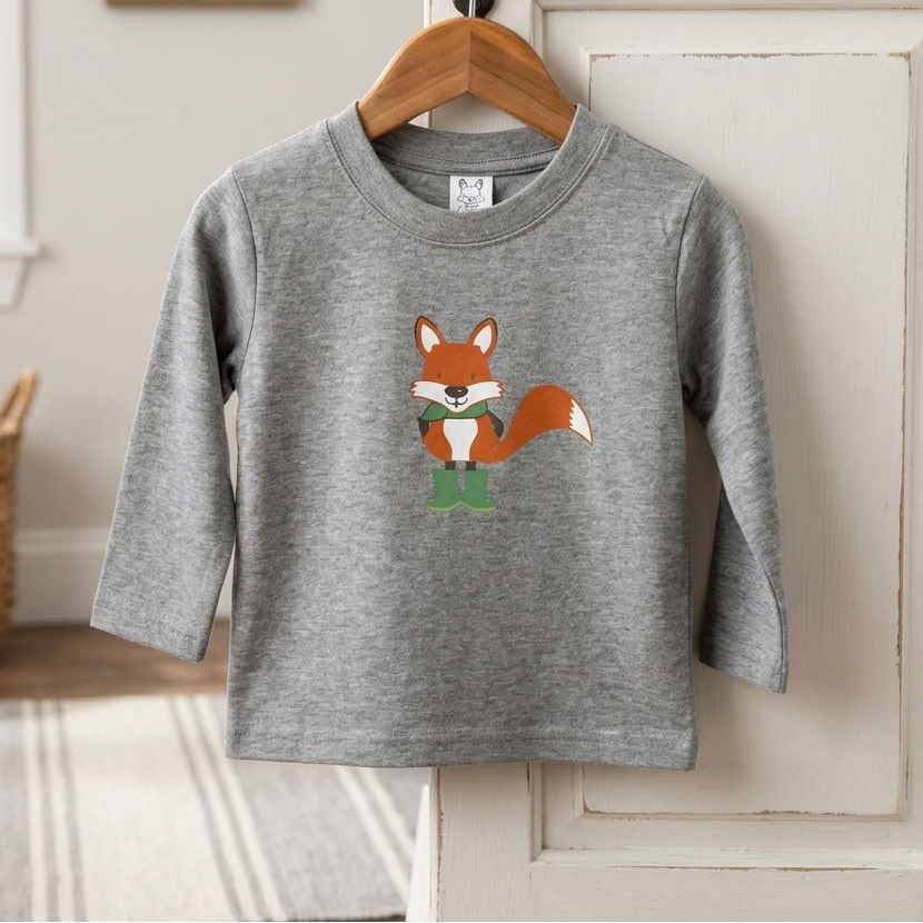 A toddler's heather gray long-sleeve shirt with a cartoon fox graphic hanging on a wooden hanger from a black hook on a white door. The background shows a cozy room setting with a basket, framed picture, and window.