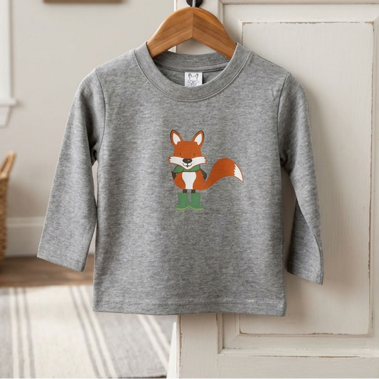 A toddler's heather gray long-sleeve shirt with a cartoon fox graphic hanging on a wooden hanger from a black hook on a white door. The background shows a cozy room setting with a basket, framed picture, and window.