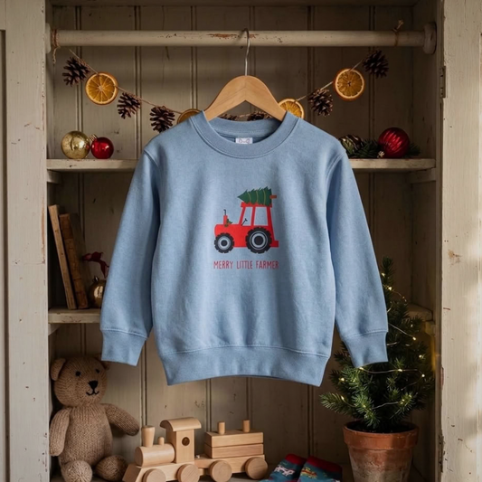 A light blue crewneck sweatshirt for kids hangs on a wooden hanger inside a rustic wooden shelf display. The sweatshirt features a graphic of a red tractor carrying a green Christmas tree, with the text 'MERRY LITTLE FARMER' printed below.