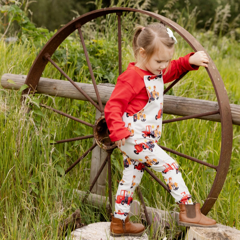 Child in a red shirt and tractor-patterned pants standing next to a large wagon wheel in a grassy area.