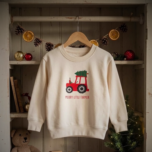 An off-white crewneck sweatshirt hanging on a wooden hanger, featuring a red tractor carrying a green Christmas tree with the text 'MERRY LITTLE FARMER' printed below. The sweatshirt is displayed on a rustic wooden shelf decorated with holiday ornaments, pinecones, dried orange slices, and a teddy bear.