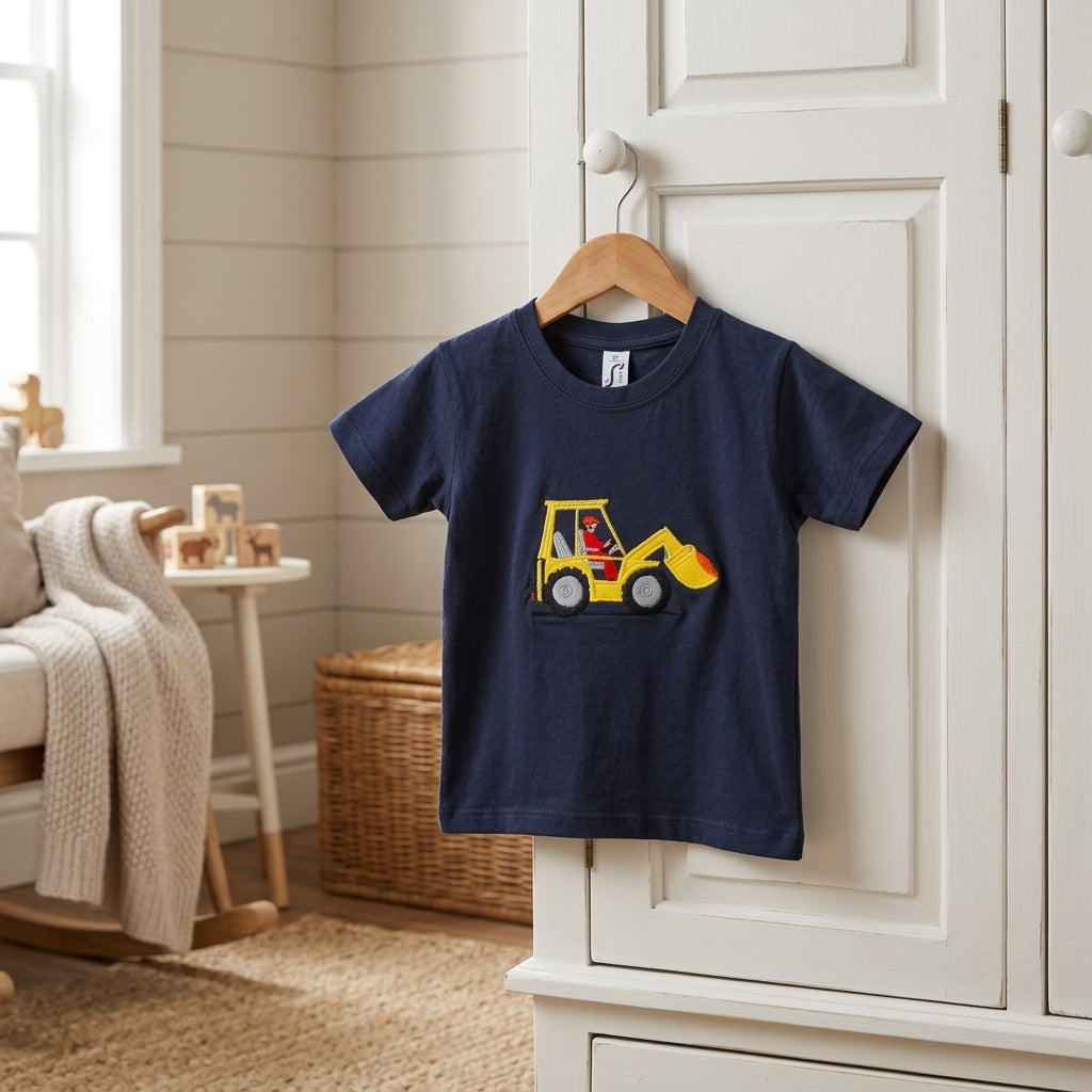 A navy blue short-sleeve kids' t-shirt hanging on a wooden hanger on a white wardrobe door in a bright, neutral nursery room. The t-shirt features a graphic of a yellow construction excavator.