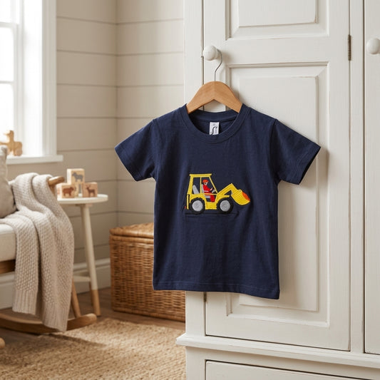 A navy blue short-sleeve kids' t-shirt hanging on a wooden hanger on a white wardrobe door in a bright, neutral nursery room. The t-shirt features a graphic of a yellow construction excavator.