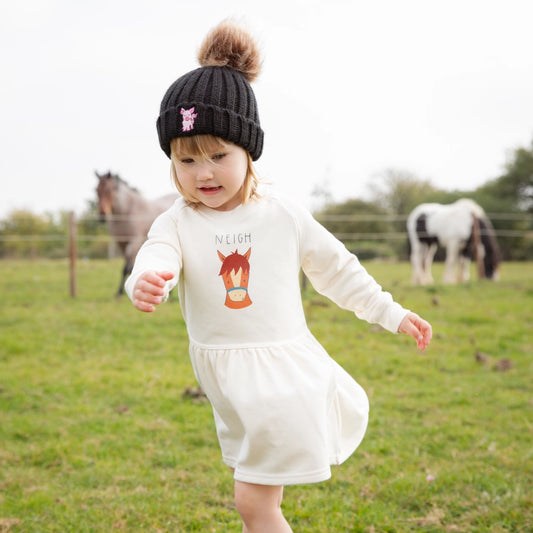 A toddler wearing a white long sleeve dress with a graphic of a horse and the word 'NIGHT' on the front, standing in a field with horses in the background.