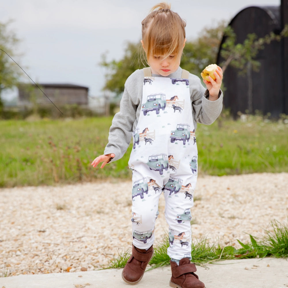 A toddler wearing white dungarees with a pattern of Land Rover vehicles, a horse, and a dog in a outdoor setting.