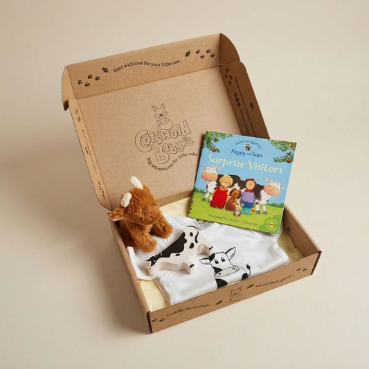 A curated children's gift box from Cotswold baby co shown open on a warm beige background. Inside, a small brown plush cow sits next to a white baby towel with a black-and-white cow print and a colorful children's book.