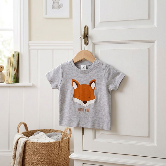 A toddler's heather gray short-sleeve t-shirt with an orange and white fox face graphic and the text 'LITTLE FOX' hanging on a wooden hanger on a white paneled door. The scene is styled with a woven basket and books in a bright, clean nursery setting.