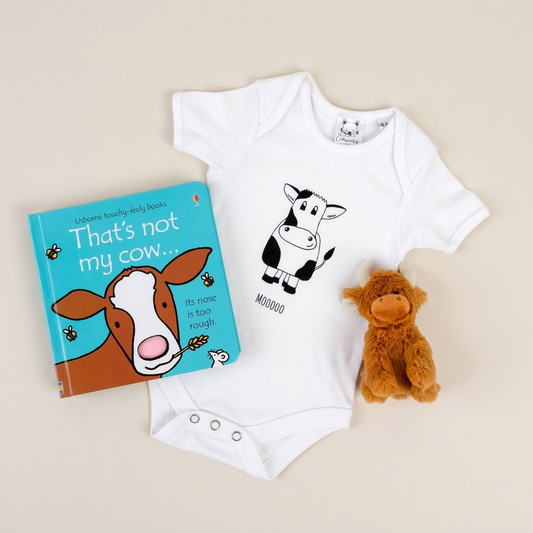 A baby bodysuit laid flat on a light beige background, styled with a children's book titled 'That's not my cow.