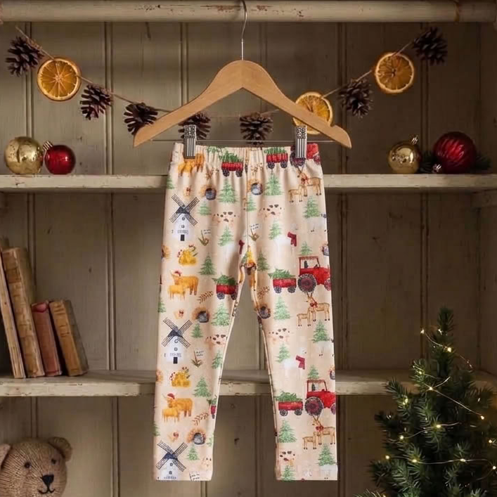 A pair of baby pants with a colorful woodland print hanging on a wooden hanger inside a rustic wooden shelf display. The pants feature a cream background with illustrations of red tractors, green pine trees, brown animals, and a blue windmill.