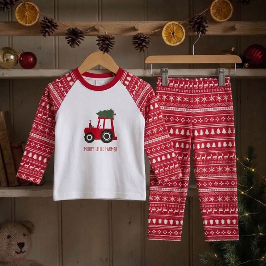 A two-piece children's pajama set hanging on wooden hangers against a rustic holiday shelf backdrop. The long-sleeve top is white on the front with red raglan sleeves and a red collar, featuring a red tractor graphic carrying a green Christmas tree and the words 'MERRY LITTLE FARMER.