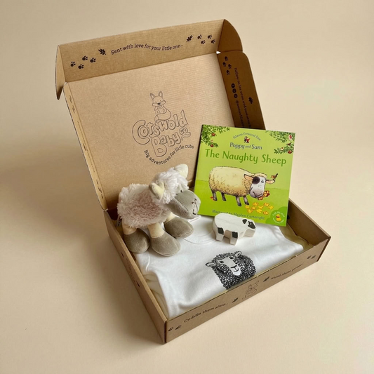 A curated children's gift box from cotswold baby co, featuring a plush sheep toy, a small white sheep figurine, a white t-shirt with a sheep print, and a children's book titled 'The Naughty Sheep' by Pepper & Sun, all arranged inside an open kraft cardboard box.