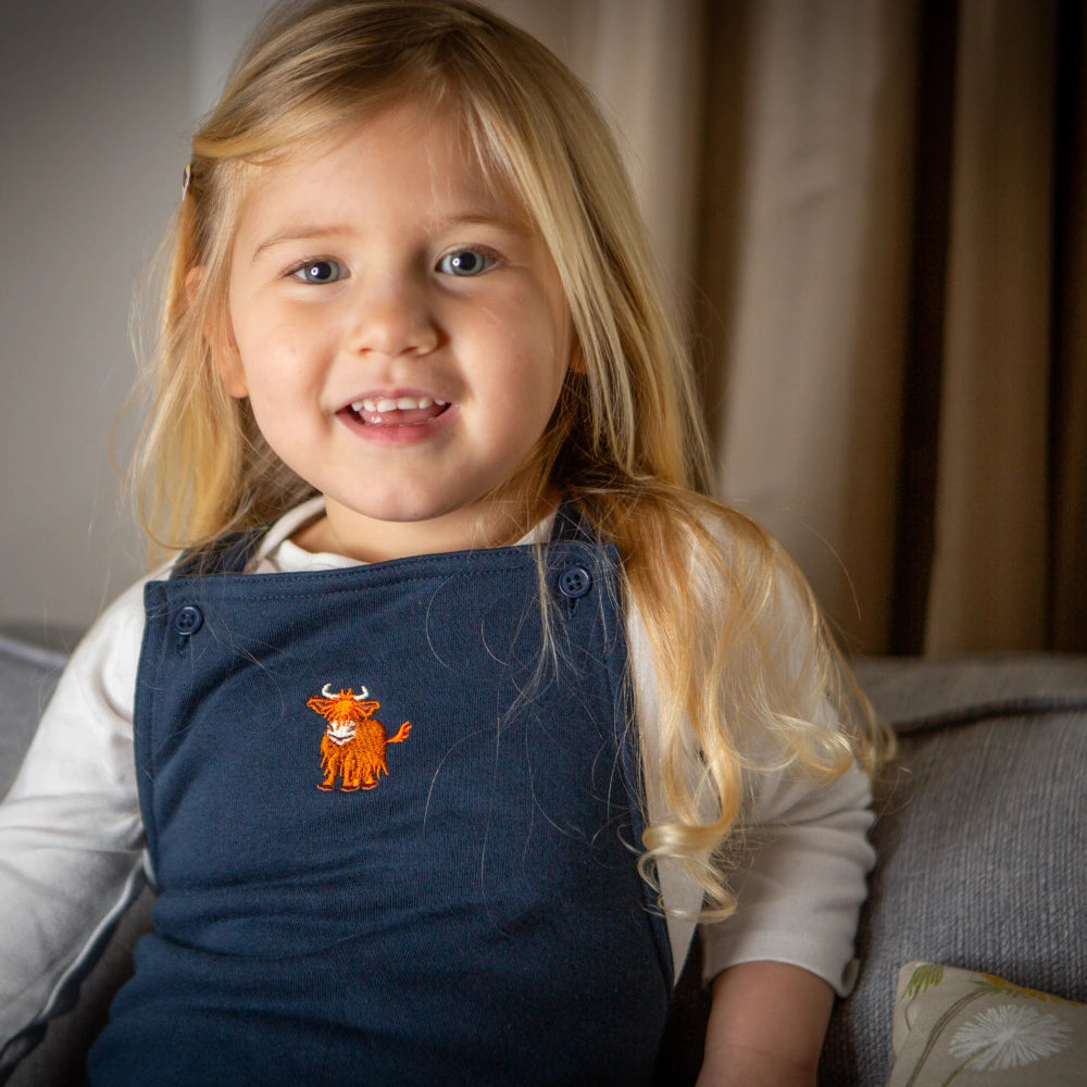 A young child wearing navy dungarees with a Highland cow embroidery on the pocket, sitting on a sofa.