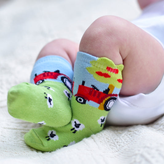 A pair of green and blue baby socks with sheep and a tractor design, displayed on a baby's feet.